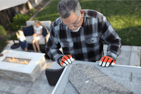 A man in safety glasses, gloves, and a plaid shirt cleans a gutter fitted with stainless steel micro-mesh. In the background, a woman and child sit by an outdoor fire pit on a patio.